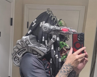 May include: A Predator-style helmet featuring long black dreadlocks with silver rings, a metallic silver shoulder cannon with a red light, and a matching silver shoulder armor piece. The helmet has green textured details and a complex mechanical design.