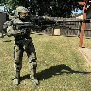 Könnte beinhalten: Ein detailliertes olivgrünes und graues Halo Spartan-Rüstungskostüm mit Helm und einer großen schwarzen und silbernen Waffe. Das Kostüm wird auf einem Rasen getragen, mit einem Holzzaun und einem Baum im Hintergrund.