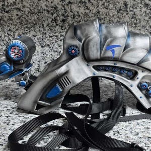 Pode incluir: Um capacete e uma arma de ficção científica prateados e azuis. O capacete tem um design curvo com detalhes azuis e uma correia preta. A arma tem um esquema de cores semelhante com detalhes intrincados.