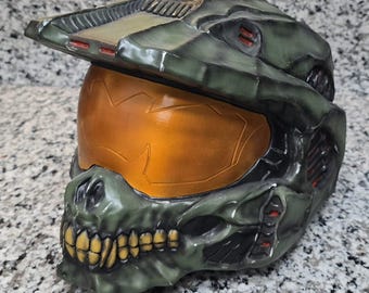 Op de afbeelding: Een gedetailleerde, groen en zwarte helm met een oranje vizier. De helm heeft een schedelachtig ontwerp op de onderkant van het gezicht en is waarschijnlijk een cosplay- of verzamelobject. De helm is gemaakt van een hard materiaal.