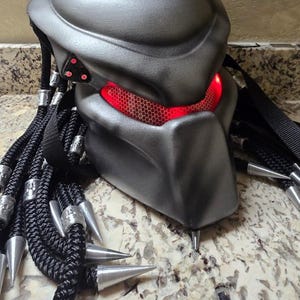 以下が含まれることがあります： 滑らかで輪郭のあるデザインのマットなダークグレーのヘルメットで、赤く光るメッシュのアイレンズが特徴です。ヘルメットの側面には3つの小さな赤いライトがあり、黒い編み込みコードが複数垂れ下がっており、それぞれに銀色の金属製のスパイクが付いています。