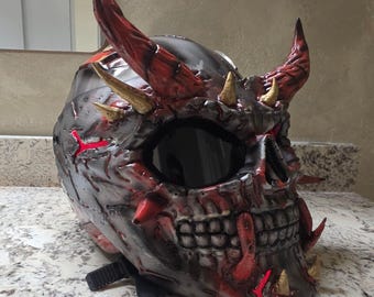Könnte beinhalten: Ein Integral-Motorradhelm mit einem individuell geformten Totenkopf-Design, großen roten Hörnern, kleineren goldenen Stacheln und einem dunklen Visier. Die Außenseite ist in verwittertem Metallic-Grau und Rot lackiert und mit roten Lichtakzenten versehen.