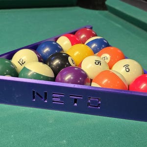Könnte beinhalten: Ein violetter, dreieckiger Billard-Ballständer mit dem Wort "NETO" auf der Vorderseite. Der Ständer ist mit 15 Billardkugeln gefüllt, jede mit einer anderen Nummer.