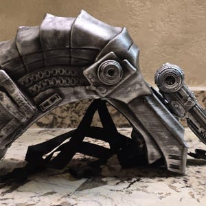 Pode incluir: Um capacete futurista prateado com detalhes intrincados e um dispositivo lateral correspondente. O capacete apresenta um design segmentado com um acabamento texturizado e um sistema de correias pretas para usar. A estética geral sugere um tema de ficção científica ou cosplay.