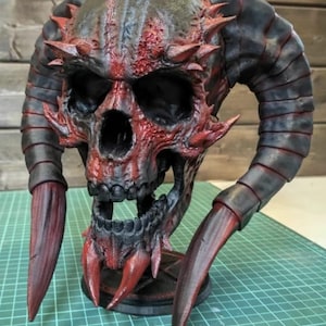 Könnte beinhalten: Eine detaillierte, 3D-gedruckte Schädel-Skulptur mit markanten Hörnern und scharfen, klauenartigen Anhängseln. Der Schädel ist in Rot- und Schwarztönen bemalt, mit strukturierten Details. Die Skulptur ist auf einem schwarzen Sockel montiert.