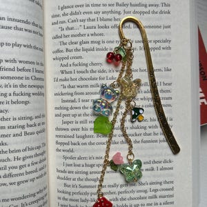 May include: Gold-toned metal bookmark with a decorative charm. The charm features red cherries, green leaves, iridescent beads, a mushroom, and a butterfly. The bookmark is placed in an open book.