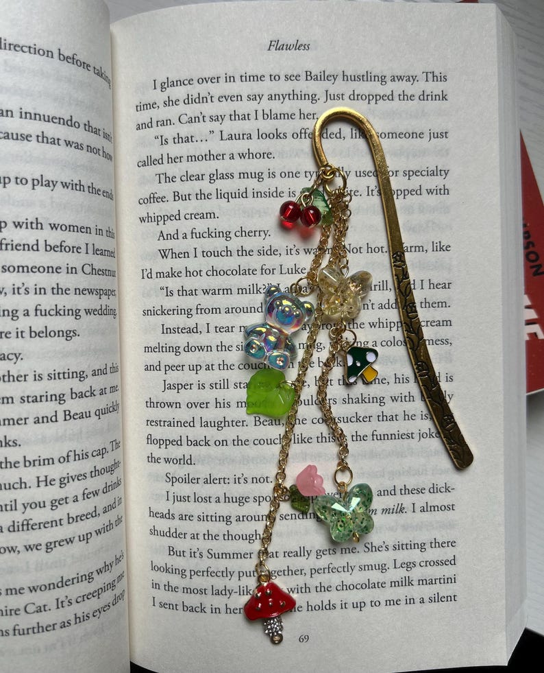 May include: Gold-toned metal bookmark with a decorative charm. The charm features red cherries, green leaves, iridescent beads, a mushroom, and a butterfly. The bookmark is placed in an open book.