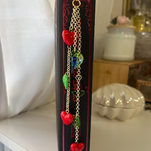 May include: A black book with red decorative accents and a gold chain bookmark. The bookmark features red heart charms, green leaf charms, and colorful glass beads. The book is on a white shelf.