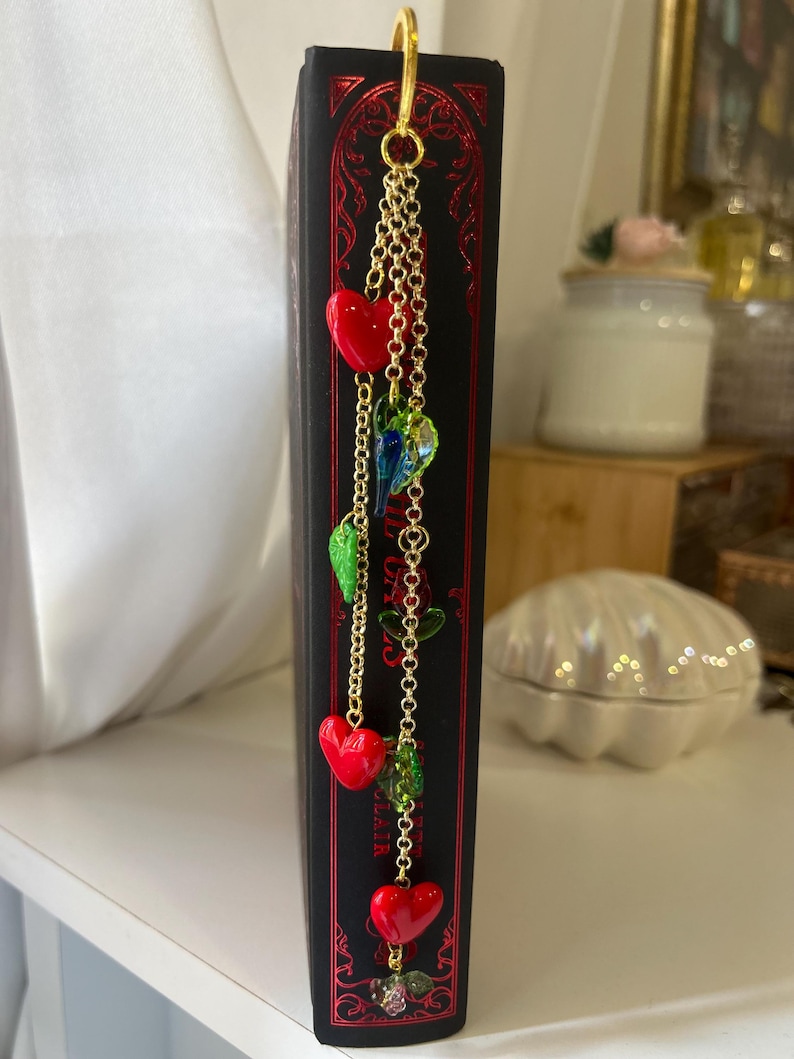 May include: A black book with red decorative accents and a gold chain bookmark. The bookmark features red heart charms, green leaf charms, and colorful glass beads. The book is on a white shelf.