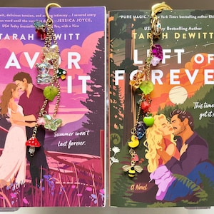 May include: Two books with colorful covers, titled "Savor It" and "Left of Forever," feature romantic illustrations. Decorative gold bookmarks with charms, including bears, butterflies, and fruit, are attached to the books. The books are novels.
