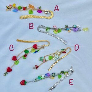May include: A collection of decorative metal bookmarks with dangling charms. The bookmarks feature various designs, including gold and silver finishes, and are adorned with colorful beads, fruit, and whimsical shapes. Each bookmark has a unique charm arrangement.