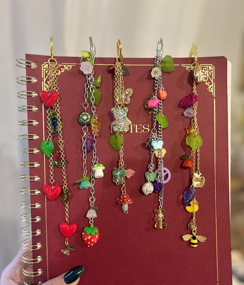 May include: Five decorative charm earrings with gold and silver chains, featuring various charms like hearts, strawberries, mushrooms, and butterflies. The earrings are displayed on a red notebook. Each earring has a different design.