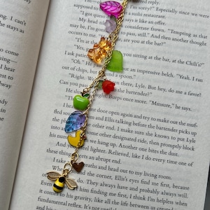 May include: A gold-toned bookmark with a chain of colorful charms, including a bee, duck, and various fruit-shaped beads. The bookmark is designed to be placed in a book, with the top portion shaped like a seashell.