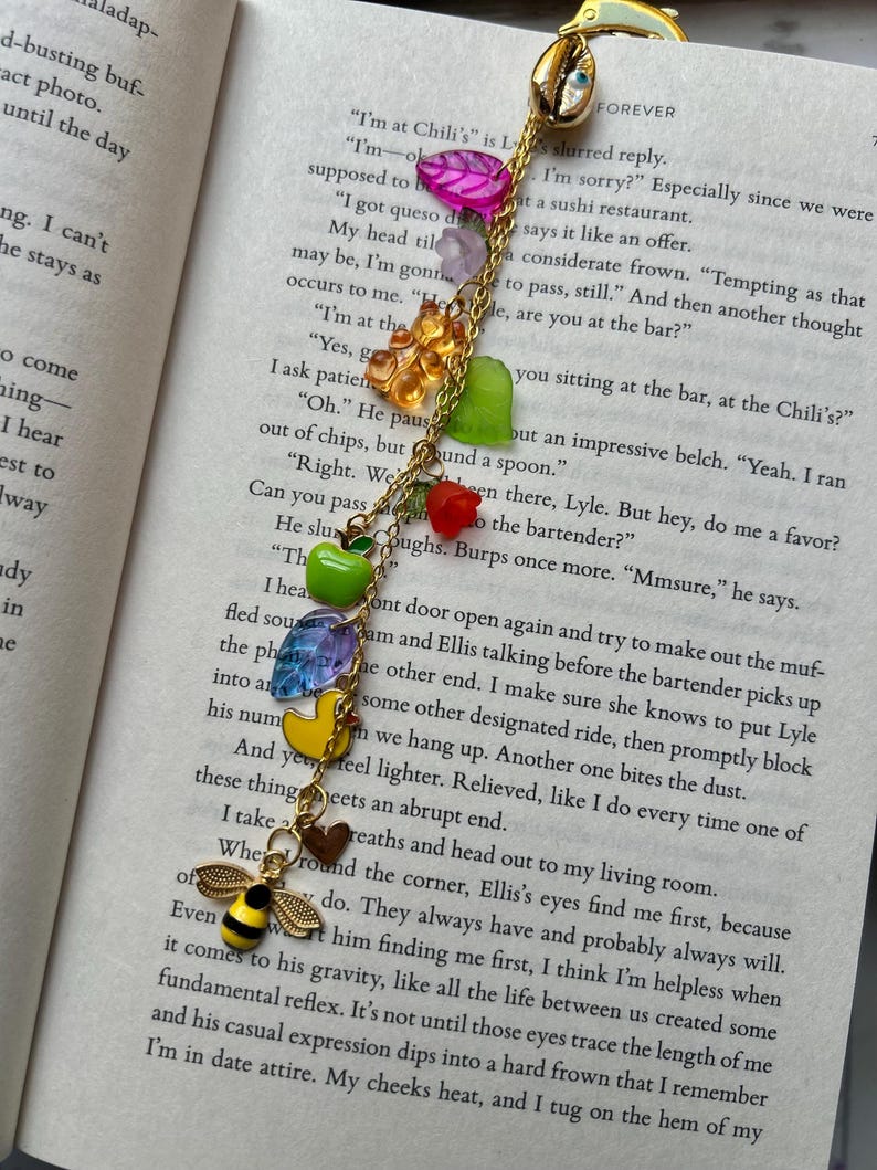 May include: A gold-toned bookmark with a chain of colorful charms, including a bee, duck, and various fruit-shaped beads. The bookmark is designed to be placed in a book, with the top portion shaped like a seashell.