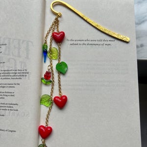 May include: A gold-colored metal bookmark with a decorative floral design. The bookmark features a chain with red heart-shaped beads, green leaf-shaped beads, and other colorful accents. Text on the book reads: "To the women who were told they must submit to the dominance of man."