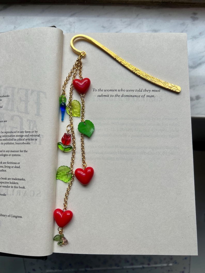 May include: A gold-colored metal bookmark with a decorative floral design. The bookmark features a chain with red heart-shaped beads, green leaf-shaped beads, and other colorful accents. Text on the book reads: "To the women who were told they must submit to the dominance of man."