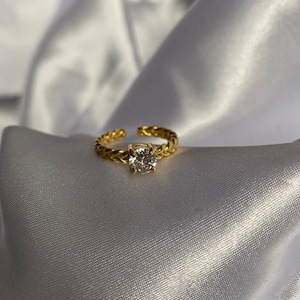 以下が含まれることがあります： 金色のリング。編み込み模様のバンドと、大きく透明な丸い宝石が特徴です。リングは下部が開いており、サイズ調整が可能です。白くシルキーな背景に置かれています。