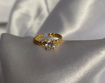 May include: A gold ring featuring a braided design and a prominent, clear, round gemstone. The ring is adjustable, with an open design at the base. It is displayed on a white, satin-like fabric.
