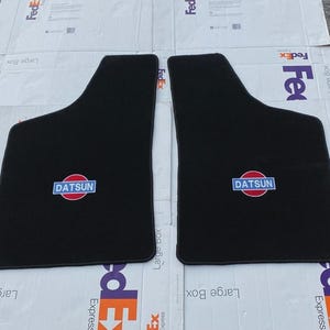 May include: A pair of black automotive floor mats featuring a Datsun logo embroidered in the center. The logo consists of a red circle with a blue rectangular banner across the middle containing the white text DATSUN in capital letters.