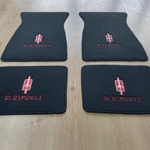 May include: A set of four black automotive floor mats featuring the Oldsmobile brand name and a red and white rocket emblem embroidered in the center of each mat. The two larger mats have a contoured shape, while the two smaller mats are rectangular.