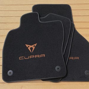 May include: A set of black automotive floor mats featuring the copper-colored Cupra logo and text embroidered on the surface. Each mat includes circular mounting grommets at the base, designed for secure placement within a vehicle interior.