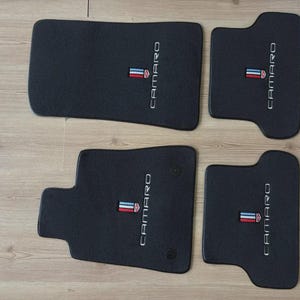 May include: A set of four black automotive floor mats featuring the word CAMARO embroidered in silver thread alongside a small rectangular emblem with red, white, and blue horizontal stripes. The mats have a textured surface and specific shapes.