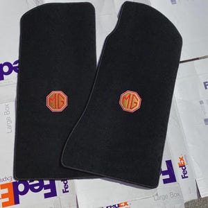 May include: Two black automotive floor mats with a textured surface. Each mat features a centered octagonal emblem with a red border and the letters MG embroidered in red thread. The mats have rounded corners and a custom contoured shape.