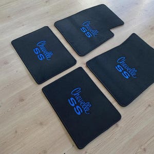May include: A set of four black automotive floor mats featuring the embroidered text Chevelle SS in bright blue thread. Each mat has a rectangular shape with rounded corners, and the mats are arranged in a scattered pattern on a light wood floor.