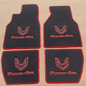 May include: A set of four black automotive floor mats with red trim. Each mat features an embroidered red Trans Am logo and a stylized bird emblem. The two front mats have an extended shape, while the two rear mats are rectangular in design.