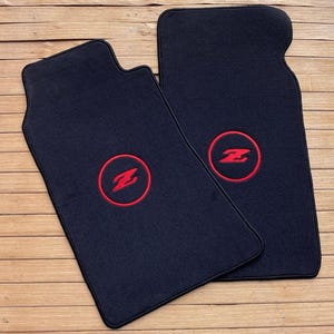 May include: Two black automotive floor mats featuring a red embroidered Z logo enclosed within a red circular border. The mats have a rectangular shape with contoured top edges and a textured surface, designed for placement in a vehicle interior.