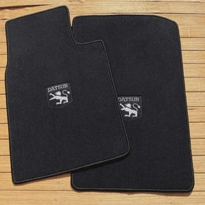 May include: Two black automotive floor mats featuring a rectangular shape with rounded corners. Each mat displays a centered embroidered patch with the text DATSUN above a white graphic of a lion standing on its hind legs with a long tail.