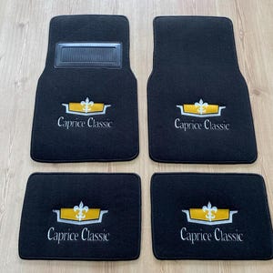 May include: A set of four black automotive floor mats featuring the embroidered text Caprice Classic and a yellow and white emblem. The two front mats include a rectangular black heel pad, while all four mats display the same logo design.