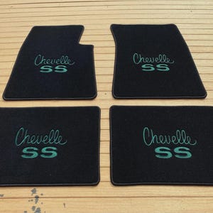 May include: A set of four black automotive floor mats featuring the text Chevelle SS embroidered in a teal script and block font. The two front mats have a notched corner design, while the two rear mats are rectangular in shape.