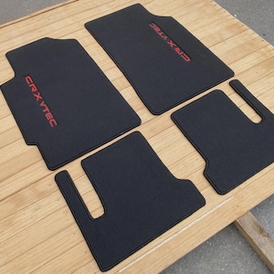 May include: A set of four black automotive floor mats with a textured surface. Two of the mats feature the red embroidered text CRX VTEC. The mats have specific cutouts and contours designed for a vehicle interior floor layout.