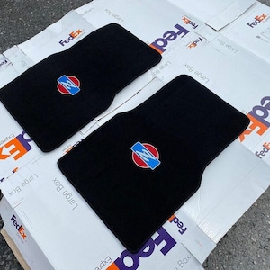May include: Two black rectangular automotive floor mats featuring a circular emblem with a red background and a blue horizontal bar containing a white letter Z. The mats have a textured carpeted surface and are shaped to fit a vehicle footwell.