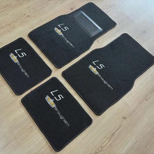 May include: A set of four black automotive floor mats featuring the embroidered text LS Brougham and a gold and silver emblem. The driver-side mat includes a rectangular textured heel pad. Each mat has a finished edge and a rectangular shape.
