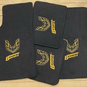 May include: A set of four black automotive floor mats featuring an embroidered yellow graphic of a stylized bird emblem and the word Bandit in a bold font. Each mat has a matching black border and a rectangular shape with rounded corners.