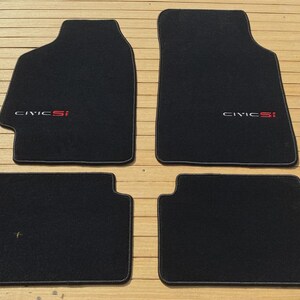 May include: A set of four black automotive floor mats with a textured surface. The two larger front mats feature the embroidered text Civic Si in white and red lettering. The two smaller rear mats are plain black with matching black trim edges.