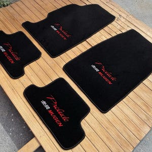 May include: A set of four black automotive floor mats with custom-shaped edges. Each mat features the text Prelude in red script and the word MUGEN in red block letters, accompanied by Japanese characters in white, embroidered on the surface.