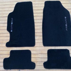 May include: A set of four black automotive floor mats with a custom shape. The two larger front mats feature the word PRELUDE embroidered in silver thread along the side. The two smaller rear mats have a matching black carpeted surface.