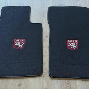 May include: Two black automotive floor mats with a rectangular shape and rounded corners. Each mat features a centered red shield emblem with white embroidery depicting a lion and the text DATSUN in a bold, capitalized white font.