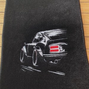 May include: A black rectangular floor mat featuring an embroidered graphic of a sports car viewed from the rear three-quarter angle. The car is depicted in white and grey thread with prominent red taillights and a visible exhaust pipe detail.