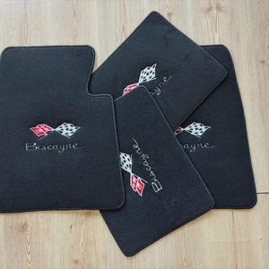 May include: A set of four black automotive floor mats featuring an embroidered logo of two crossed checkered flags in red, white, and black, with the word Biscayne stitched in white thread below the flags on each individual mat.