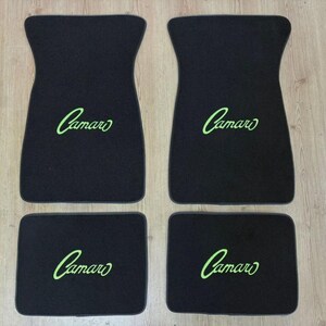 May include: A set of four black automotive floor mats featuring the word Camaro embroidered in bright green script. The set includes two larger front mats with a contoured shape and two smaller rectangular rear mats, all with matching text.