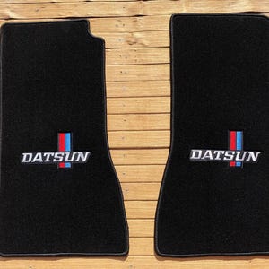 May include: A pair of black rectangular automotive floor mats with rounded corners. Each mat features the white text DATSUN embroidered above a vertical red and blue stripe emblem centered on the lower portion of the carpeted surface.