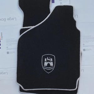 May include: A set of two black automotive floor mats with a light gray piped edge. Each mat features a centered white embroidered crest depicting a castle tower above wavy lines. The mats have a contoured shape designed to fit a vehicle floor.