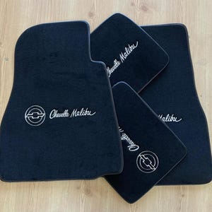 May include: A set of four black automotive floor mats featuring the white embroidered text Chevelle Malibu and a circular Chevrolet bowtie logo. The mats have a soft, carpeted texture and are arranged in an overlapping pattern on a light surface.