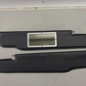 May include: Two black automotive sun visors with a smooth, leather-like texture. One visor features a rectangular vanity mirror framed in silver-toned metal, secured with four small screws. Both visors include metal mounting pins on the sides.