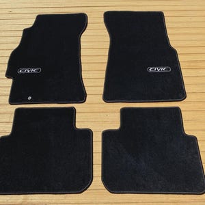 May include: A set of four black automotive floor mats with a textured surface. The two front mats feature the word CIVIC embroidered in white lettering within a rectangular border. Each mat has a distinct shape designed to fit a vehicle interior.