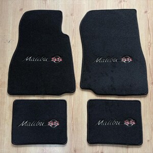 May include: A set of four black automotive floor mats featuring the embroidered text Malibu SS in silver and red. The two larger front mats have a contoured shape, while the two smaller rear mats are rectangular, all with a carpeted texture.